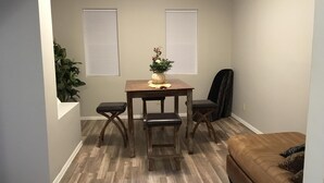 Un coin repas avec une table en bois, quatre chaises et un vase de fleurs. Il y a deux fenêtres équipées de stores, un canapé marron et une plante dans un coin.