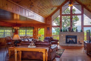 A spacious living room with a fireplace, leather sofas, wooden furniture, and large windows offering a view of the outdoors. - MountainViews: Views, Hot Tub, Wifi, Resort Pool, near Dollywood & Pigeon Forge (Sevierville)