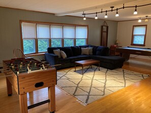 A living room with a foosball table, a dark-colored sectional sofa, a pool table, and large windows with blinds.