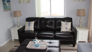 A living room with a black leather sofa, two white nightstands with lamps, a window with a view, and decorative pillows. - Magnificent Bay View!!! Luxurious Condo, Your Home-Away-From-Home, Makai #316 (Ocean City)