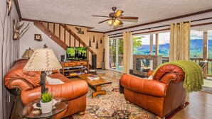A spacious living room with leather sofas, a wooden coffee table, and a television. A ceiling fan and a fireplace are visible, along with a large window offering a scenic view. - 🐻Awesome View: Mtn Views! Secluded, Hot Tub, WiFi, Near Attractions!🗻 (Sevierville)