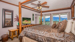 A spacious bedroom with a large bed, wooden ceiling beams, a ceiling fan, a television, and a scenic view through the windows. - 🐻Awesome View: Mtn Views! Secluded, Hot Tub, WiFi, Near Attractions!🗻 (Sevierville)