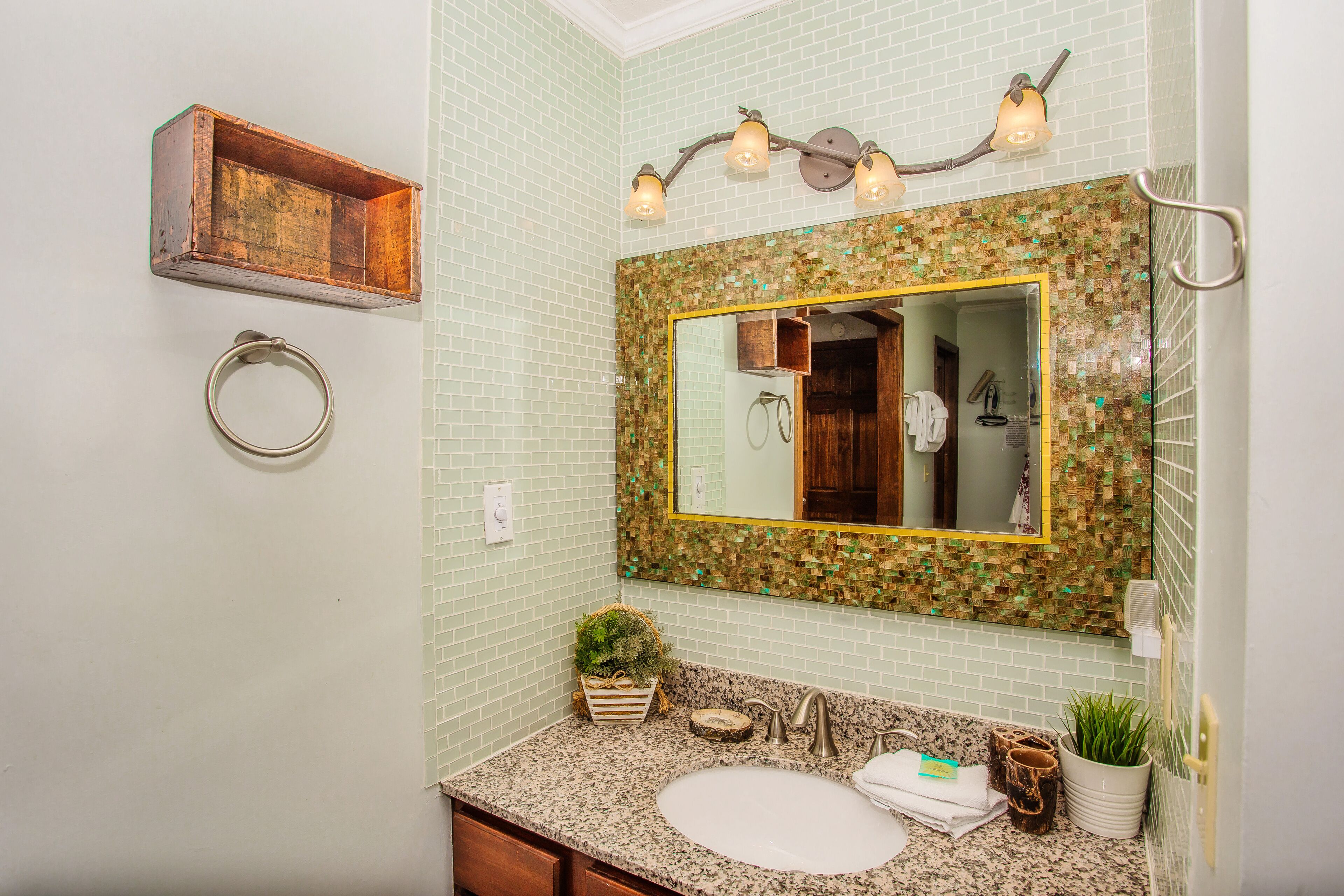 Une salle de bain dotée d’un comptoir en granit, d’un lavabo, d’un miroir avec un pare-feu en mosaïque, d’un porte-serviettes et d’une étagère en bois.
