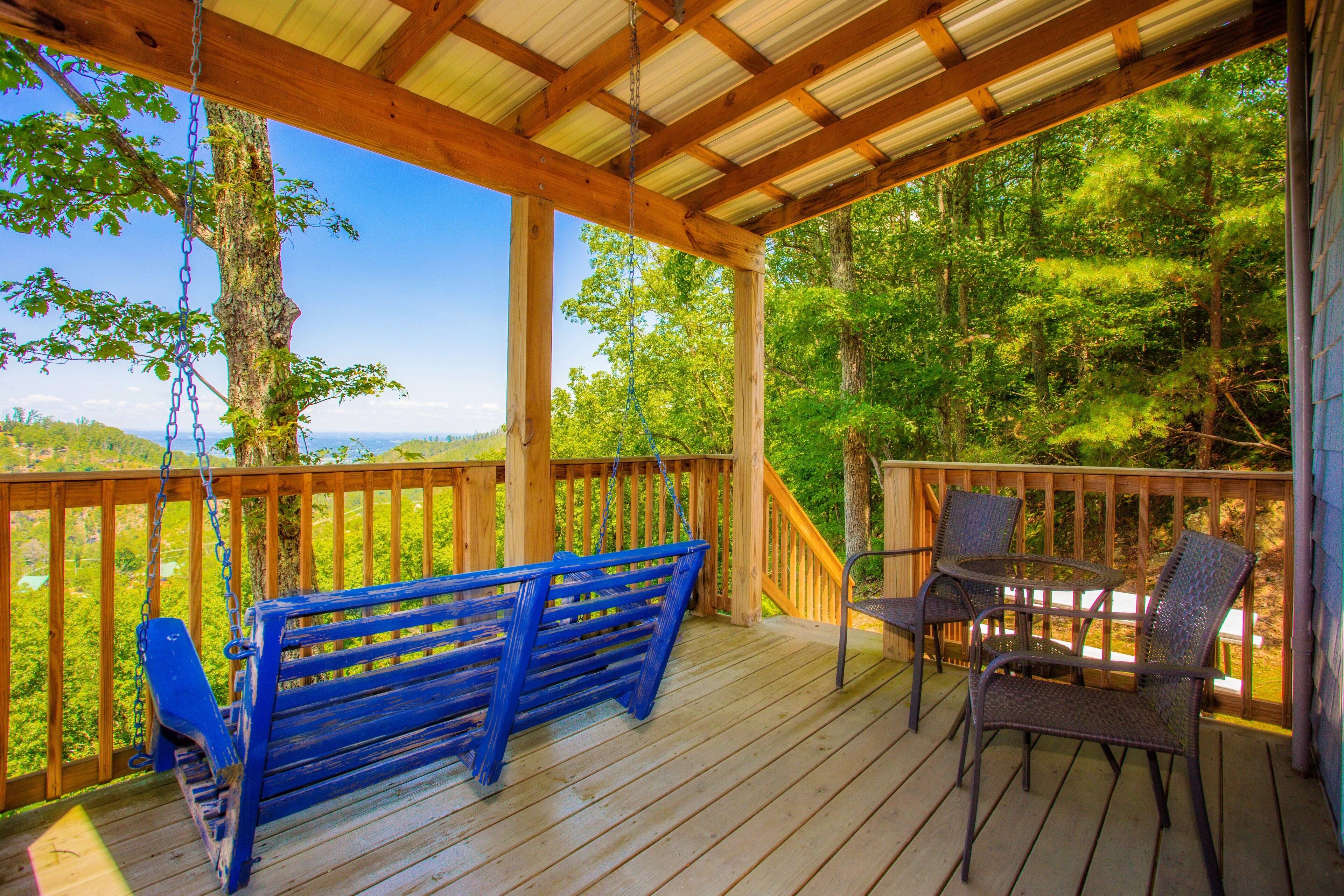 A wooden deck with a swing set and metal chairs, overlooking a forested area.
