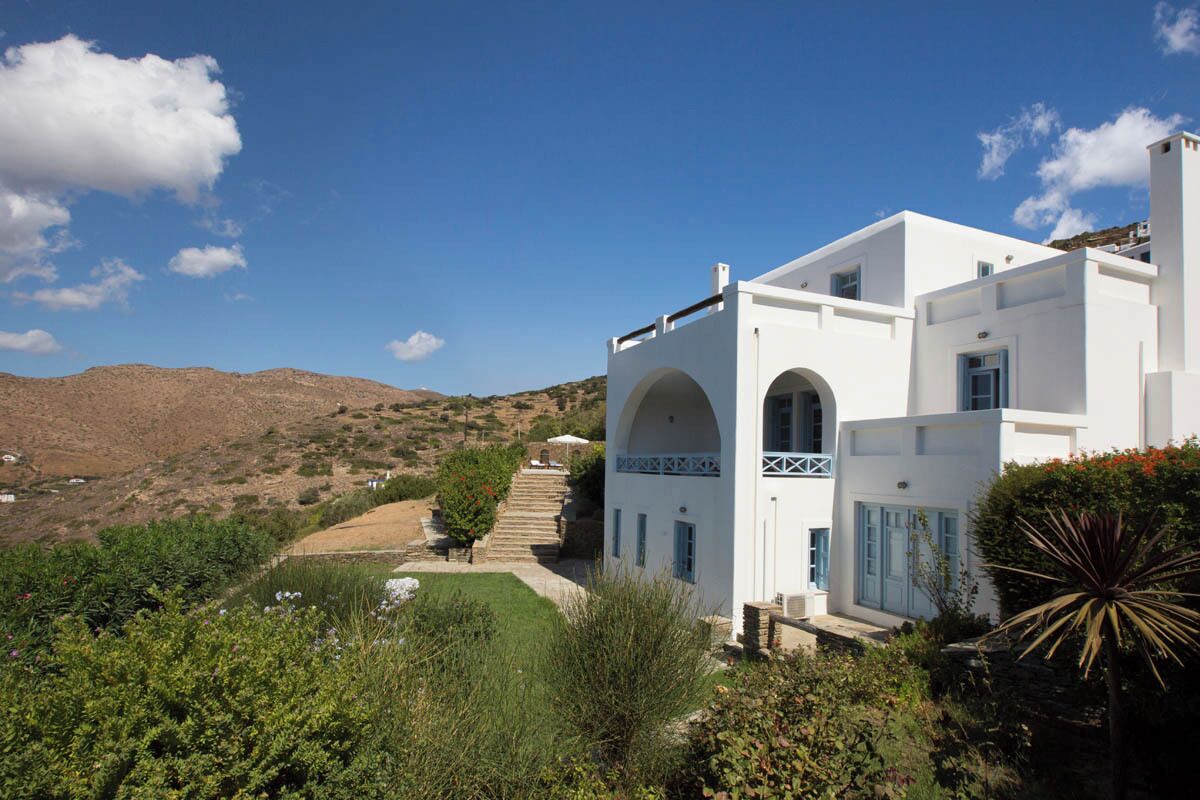 A white building with arched windows and a balcony, surrounded by greenery and a hillside.