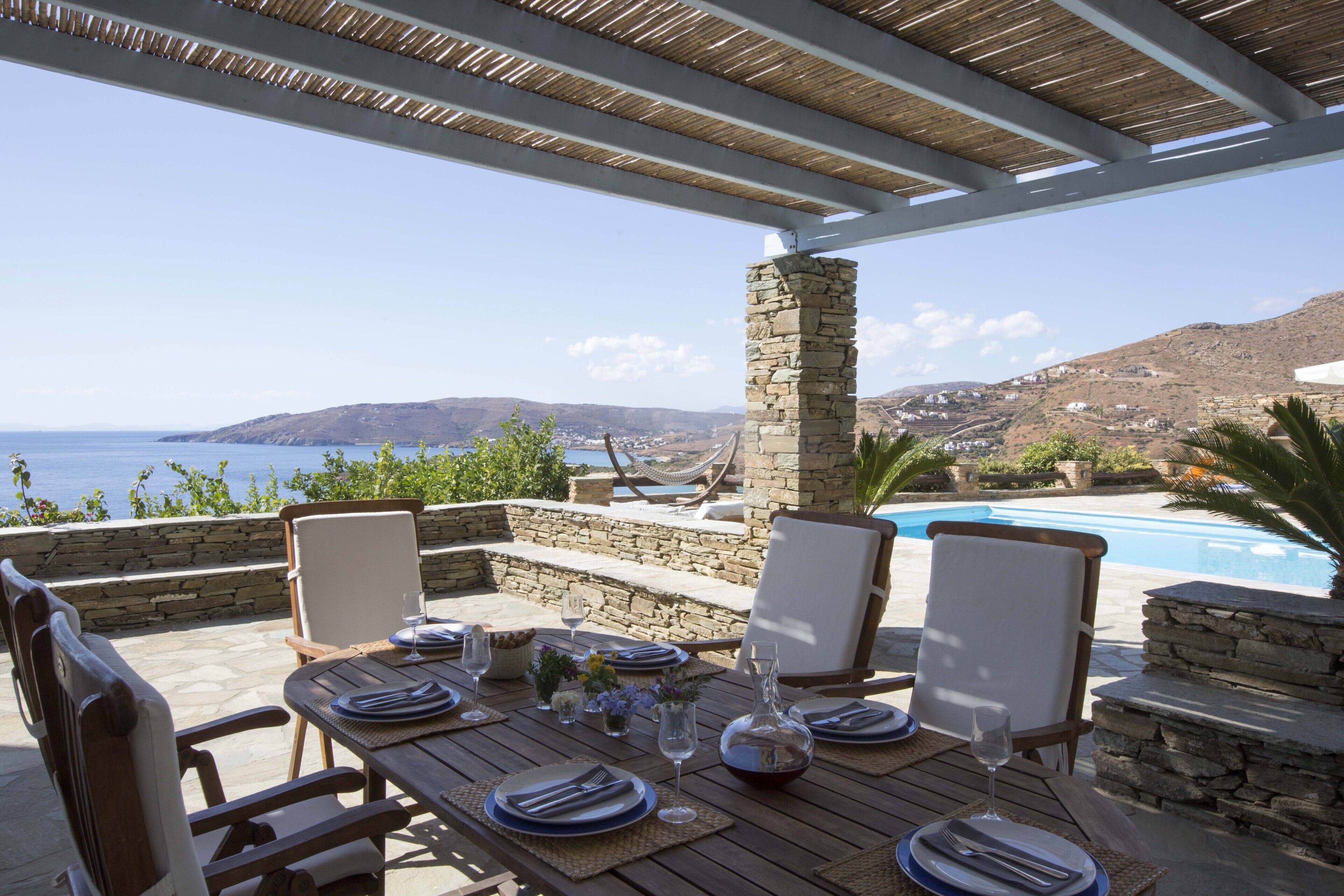 An outdoor dining area with a table set for four, overlooking a pool and a scenic view of the sea and hills.