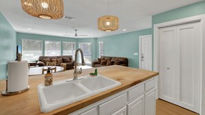A modern kitchen with a wooden countertop, white cabinets, and a double sink. There is a ceiling fan, two pendant lights, and a view of a living room with a couch and a window. - Riviera | Pools & Hot-Tubs | Pet-Friendly* | King Bed (New Braunfels)