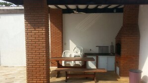 Uma área de varanda coberta com um pilar de tijolo, uma mesa de madeira, cadeiras de plástico branco, uma piscina e uma churrasqueira.