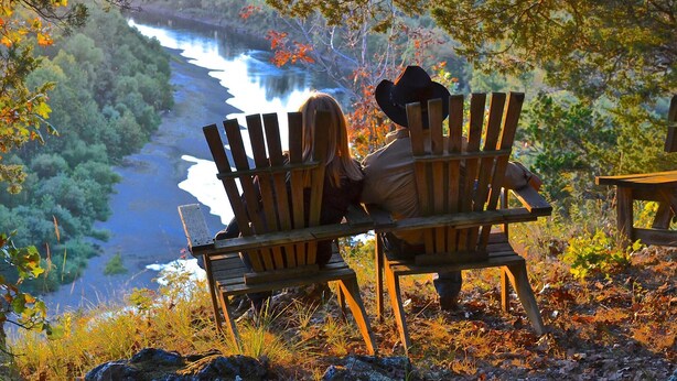 To træstole af Adirondack-design vendt mod et naturskønt udsigtspunkt over en flod og en skov.