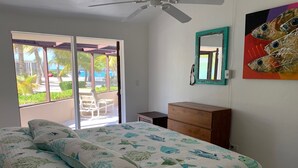 A bedroom with a bed, a wooden dresser, a ceiling fan, and a window with a view of palm trees and a pool.