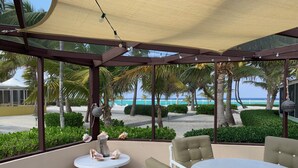 A covered outdoor seating area with a table and chairs, overlooking a beach with palm trees and the ocean.