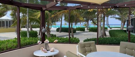 A covered outdoor seating area with a table and chairs, overlooking a beach with palm trees and the ocean.