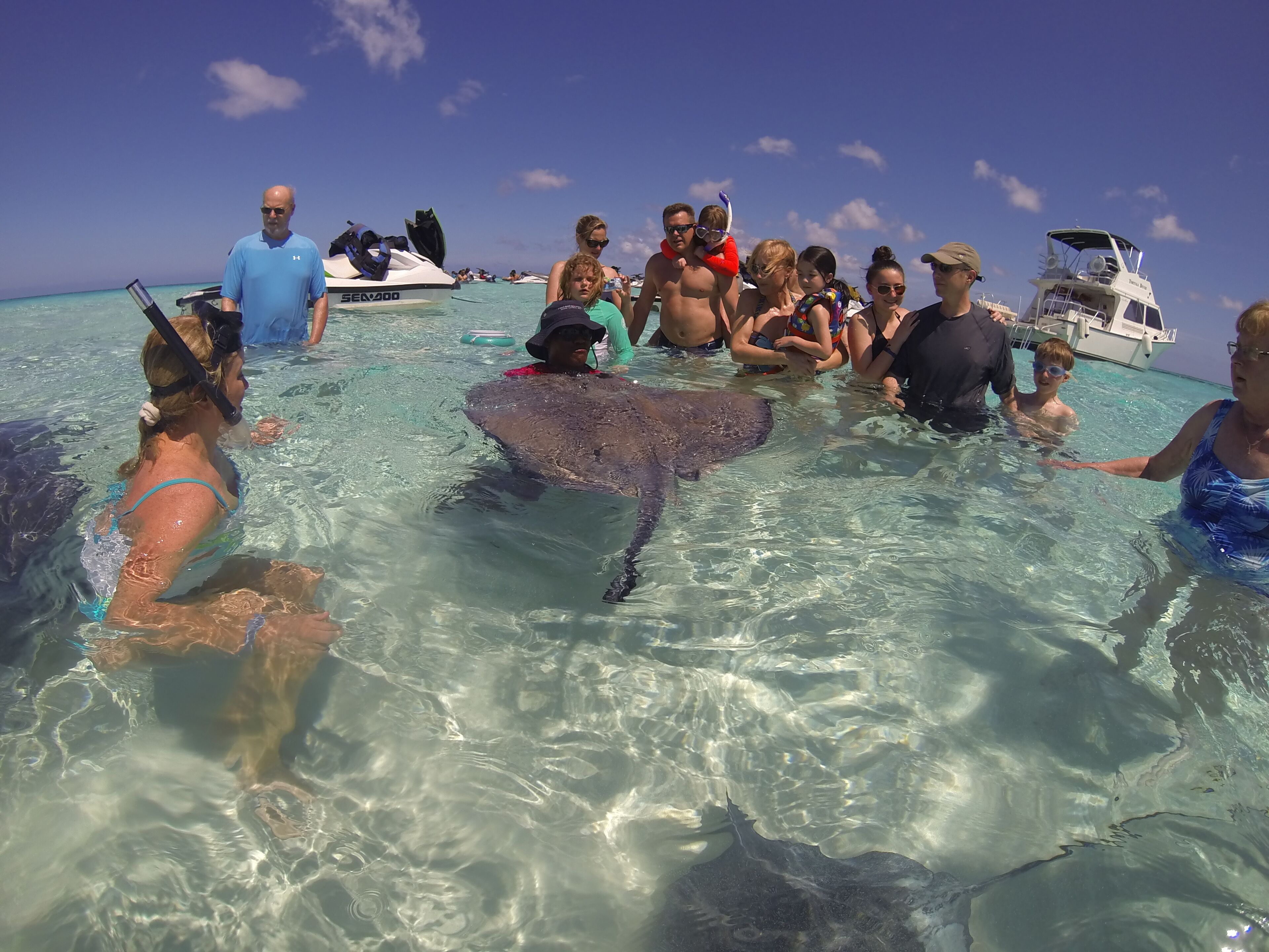 A group of people in shallow water with a stingray, boats, and a clear blue sky.