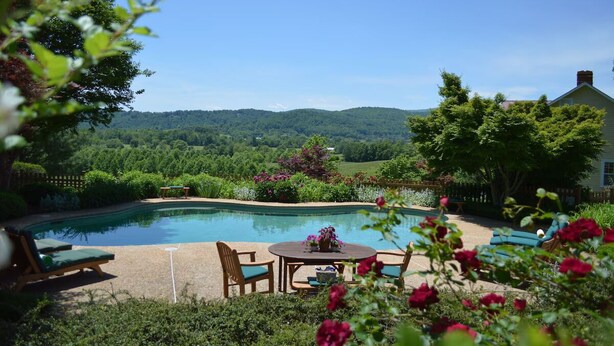 Une piscine avec vue sur une colline boisée, une terrasse avec une table et des chaises, et une maison en arrière-plan.