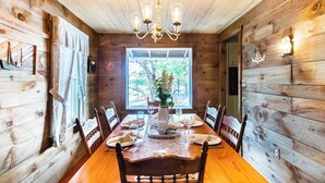 A wooden cabin interior with a dining table set for four, a chandelier, and a large window with a view of trees. - Waterfront Views of Creek- Beach Access! (Wellfleet)