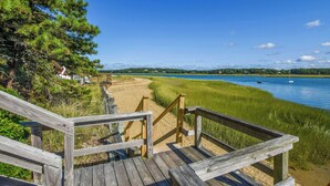 A wooden boardwalk leading to a beach with a clear view of a lake and distant houses. - Waterfront Views of Creek- Beach Access! (Wellfleet)