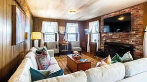 A cozy living room with a fireplace, wooden walls, and a flat-screen TV mounted on the brick wall. - Waterfront Views of Creek- Beach Access! (Wellfleet)