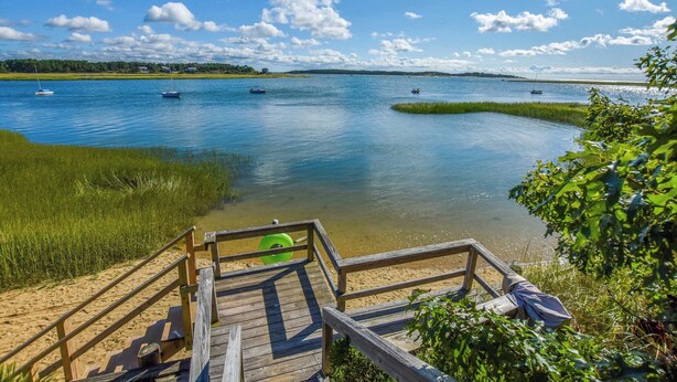 A wooden deck with a green inflatable ring, a sandy beach, and a calm lake with boats. - Waterfront Views of Creek- Beach Access! (Wellfleet)