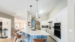 Una cocina moderna con armarios blancos, una isla grande con taburetes de bar, electrodomésticos de acero inoxidable e iluminación colgante.