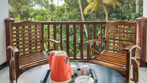 A wooden bench and table with a tray containing a red kettle, cups, and a spoon, set against a backdrop of lush greenery and palm trees. - Ashiyana (Mirissa)