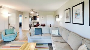 A modern living room with a sofa, a coffee table, and a colorful rug. There is a kitchen in the background with a dining area.
