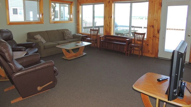 A living room with a sofa, chairs, a coffee table, a TV, and a balcony with wooden railings. - Cottage #10 - 3 bedroom/ 1 bath (Cape Vincent)