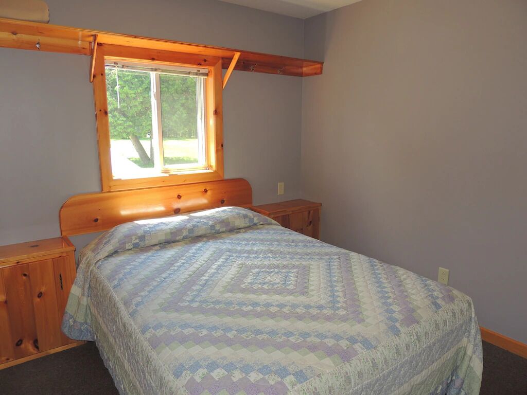 A bedroom with a wooden headboard, a window with a view of trees, a bed with a patterned quilt, and a wooden nightstand.