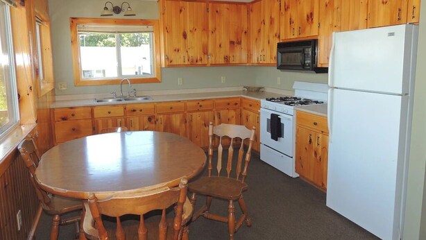 A kitchen with wooden cabinets, a stove, a microwave, a refrigerator, a dining table with chairs, and a ceiling fan. - Cottage #10 - 3 bedroom/ 1 bath (Cape Vincent)