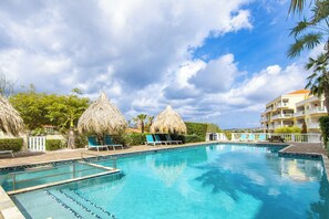 A swimming pool with lounge chairs and thatched umbrellas, surrounded by greenery and a building with balconies.
