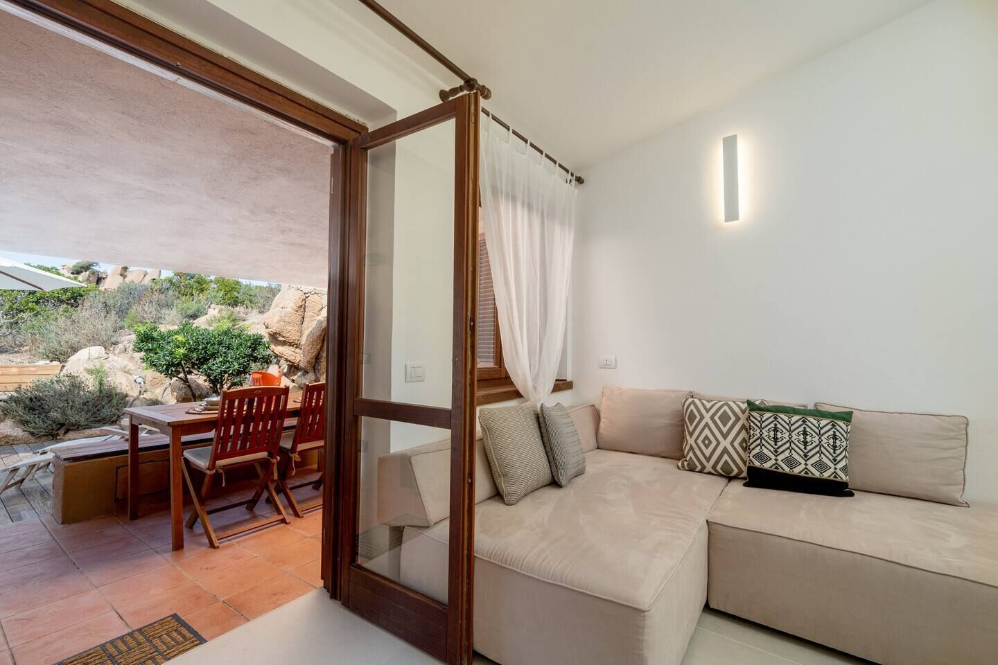 A cozy living area with a beige sofa, a wooden dining table, and chairs. A glass door leads to an outdoor patio with a table and chairs.