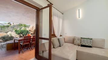 A cozy living area with a beige sofa, a wooden dining table, and chairs. A glass door leads to an outdoor patio with a table and chairs.