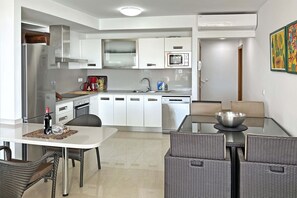 A modern kitchen with white cabinets, a stainless steel refrigerator, and a microwave. There is a dining area with wicker chairs and a glass table. - Villa Salobre Golfers 1 (Maspalomas)