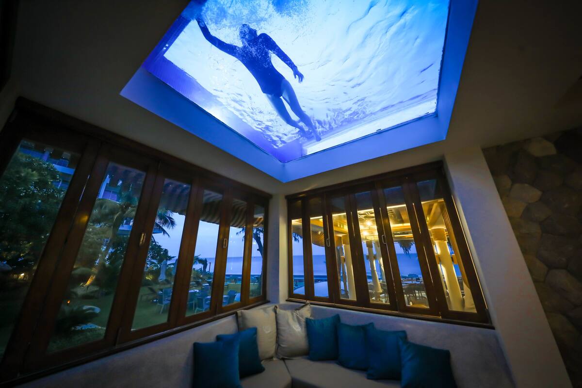 A room with a ceiling projection of a person swimming, large windows with a view of the sea, and a seating area with cushions.