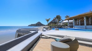 Zona de piscina en un resort, con vista a la playa y al océano, sillas de descanso y una gran formación rocosa al fondo.