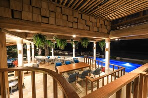 Uma área externa com mesas e cadeiras de madeira, piscina e plantas penduradas.