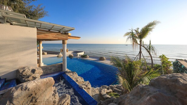 Zona de piscina con vista al mar, rodeada de rocas y palmeras.