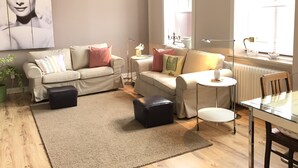 A living room with a beige sofa, a coffee table, and a dining table with chairs. There is a large window with blinds, a floor lamp, and a potted plant. - Charming, spacious town house, Friedrichstadt n. North Sea / 2-4 pers (Friedrichstadt)