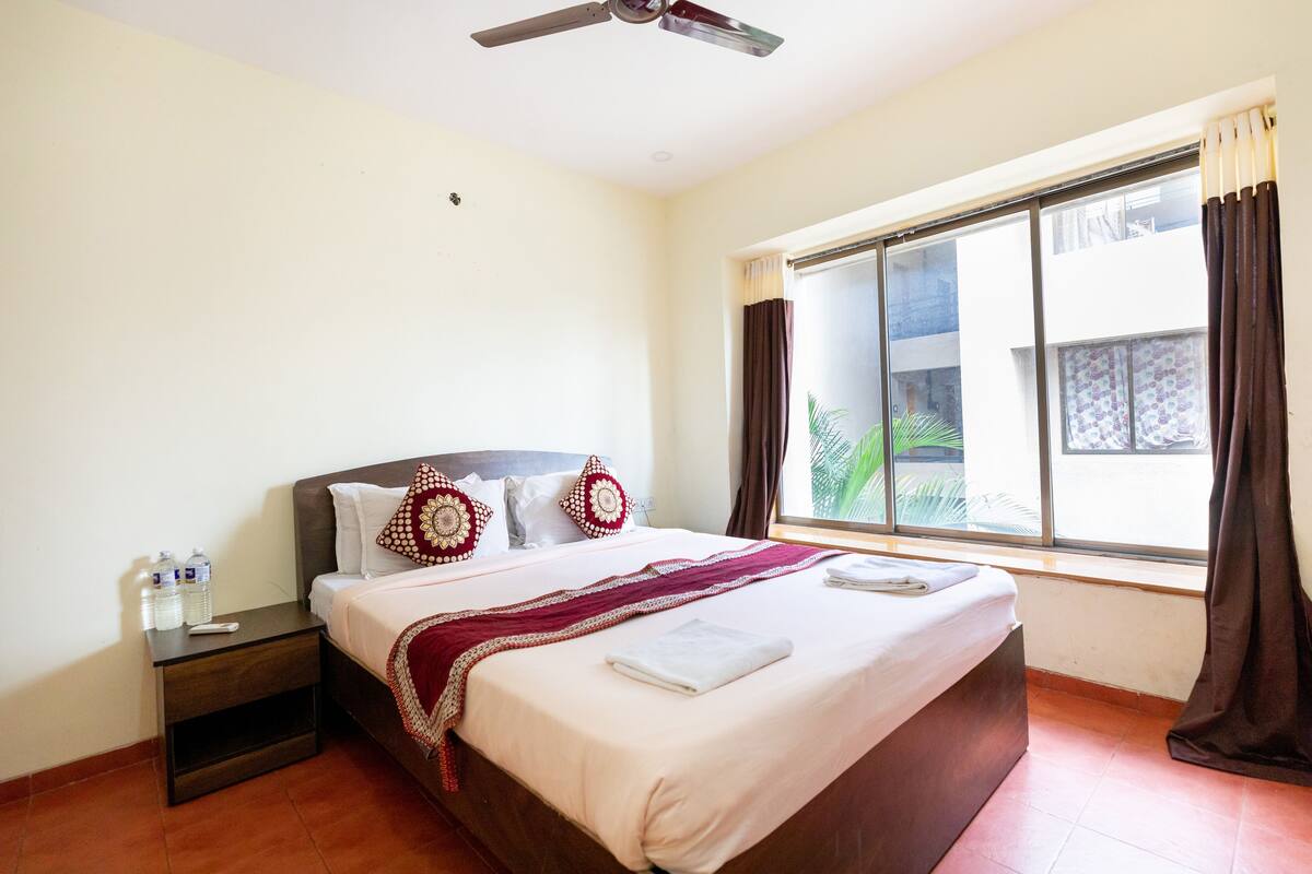 Quarto de hotel com cama, mesa de cabeceira, ventilador de teto e uma janela grande com cortinas.