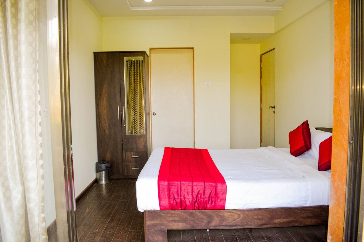 Quarto de hotel com estrutura de cama de madeira, colcha branca e almofadas vermelhas. Há um guarda-roupa de madeira e uma porta com cortina.