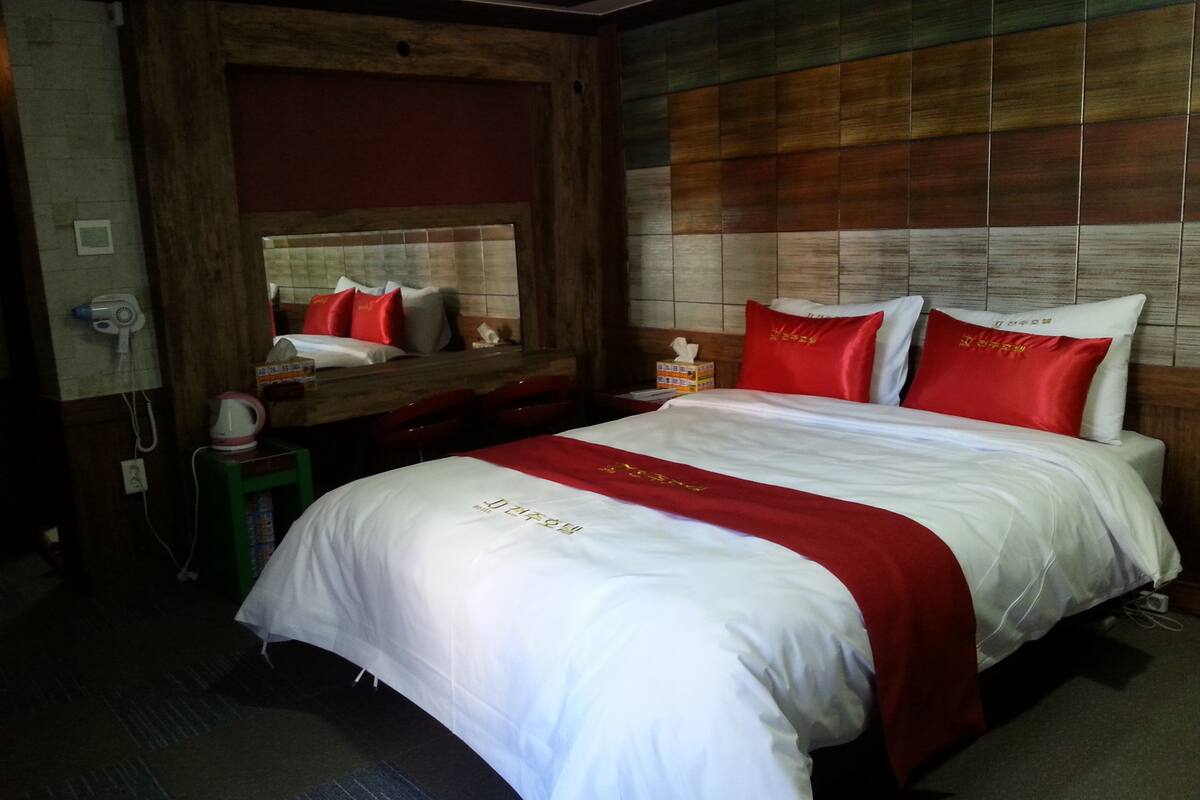 A hotel room with a large bed, a wooden headboard, a bedside table with a lamp, and a small stool.