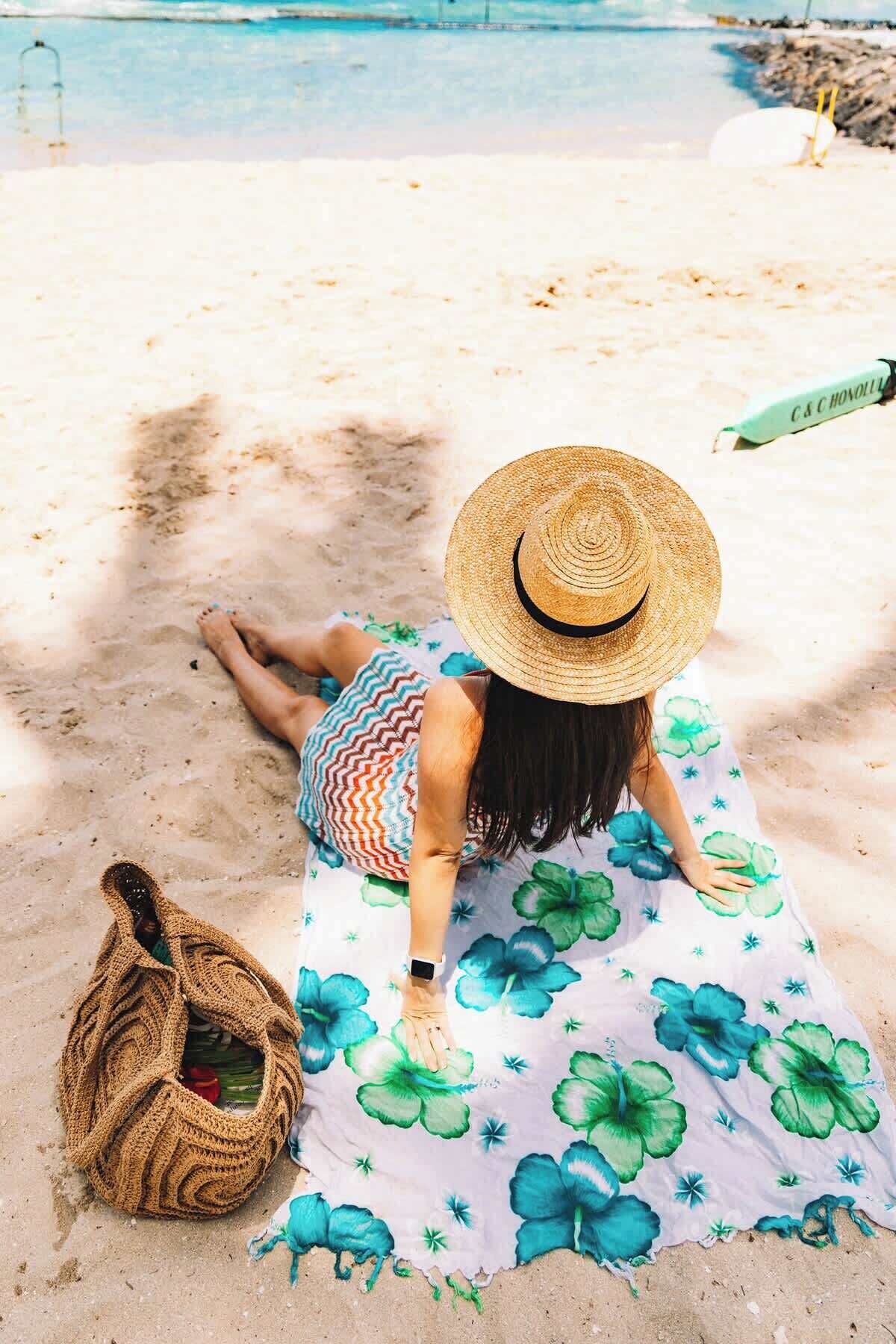 Una persona recostada sobre una toalla de playa con estampado floral, un sombrero de paja y un bolso tejido.
