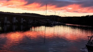 En havn med fortøjede både, roligt vand der spejler solnedgangen, og en bro i baggrunden.