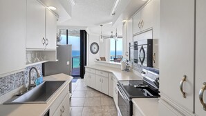 A modern kitchen with white cabinetry, a stainless steel sink, and a view of the ocean through an open door. - Surfside 1501: Gulf Front Luxury Lookout (Miramar Beach)
