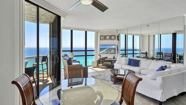A spacious living area with a large glass table, comfortable seating, and a ceiling fan. A balcony with a view of the ocean is visible. - Surfside 1501: Gulf Front Luxury Lookout (Miramar Beach)