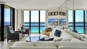 A modern living room with a large window overlooking the ocean, a white sofa, a coffee table, and a fireplace. - Surfside 1501: Gulf Front Luxury Lookout (Miramar Beach)