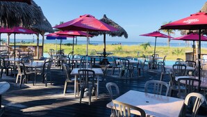 An outdoor dining area with tables and chairs, red umbrellas, and a view of the beach. - Bayside Condo with Brand New Interior, Walkable to BEACH, Retail, Dining & more! (Madeira Beach)