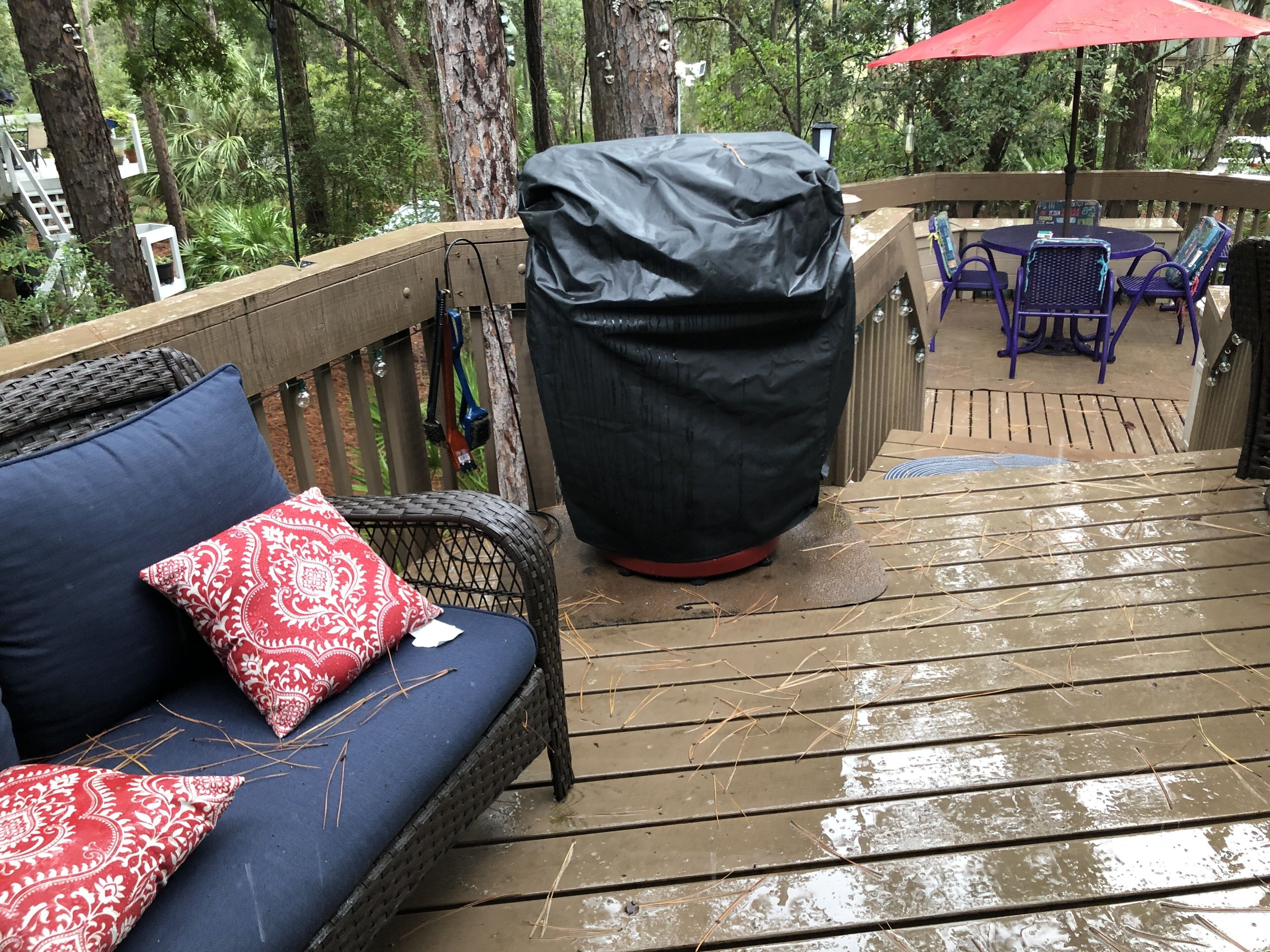 En træterrasse med grill under tag, en fletsofa med røde puder og et bord med stole.