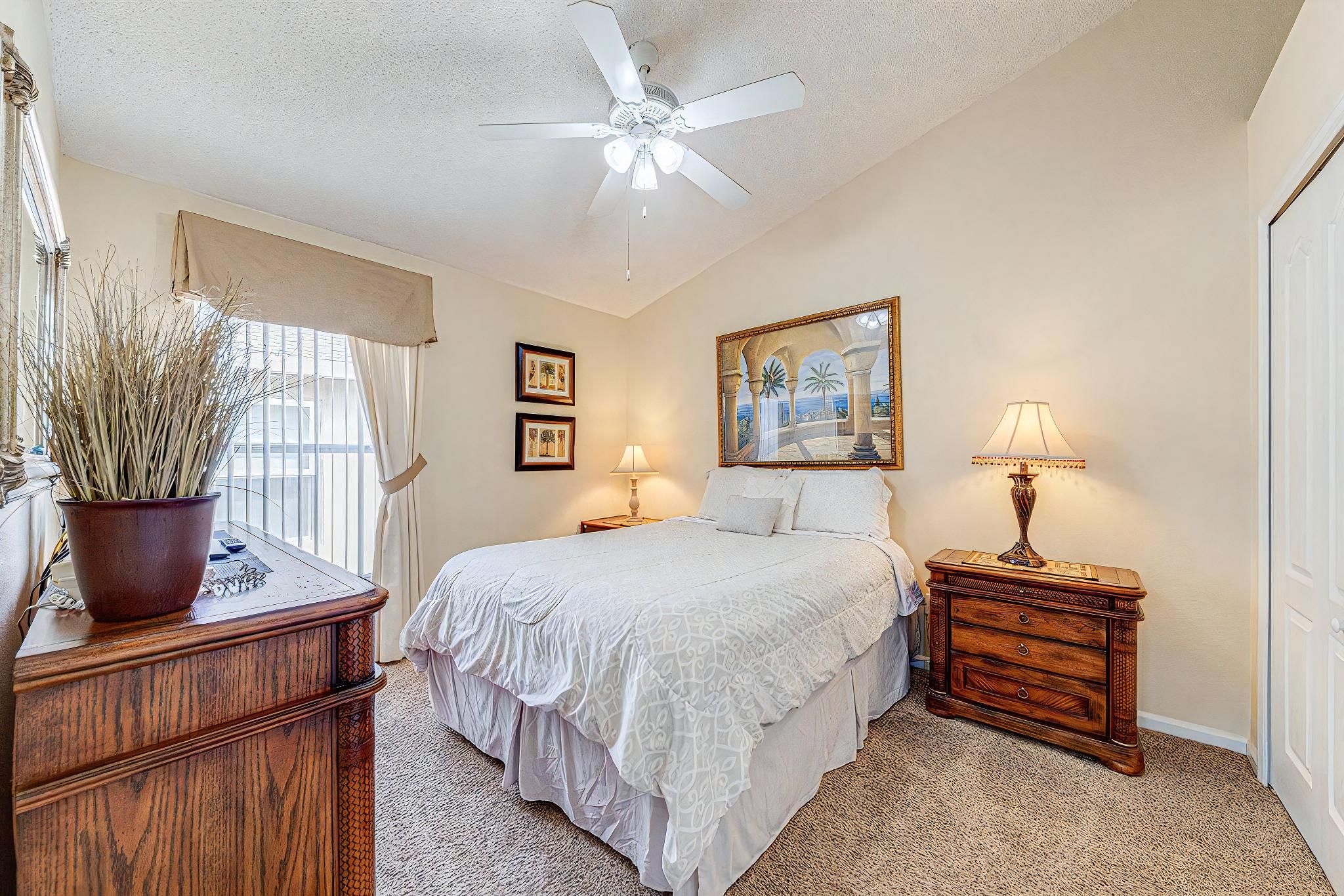 A bedroom with a bed, a ceiling fan, a nightstand with a lamp, a dresser, and a window with curtains.