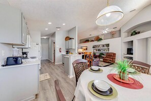 A modern kitchen with white cabinets, a dining area with a round table, and a living room with a sofa and a TV.