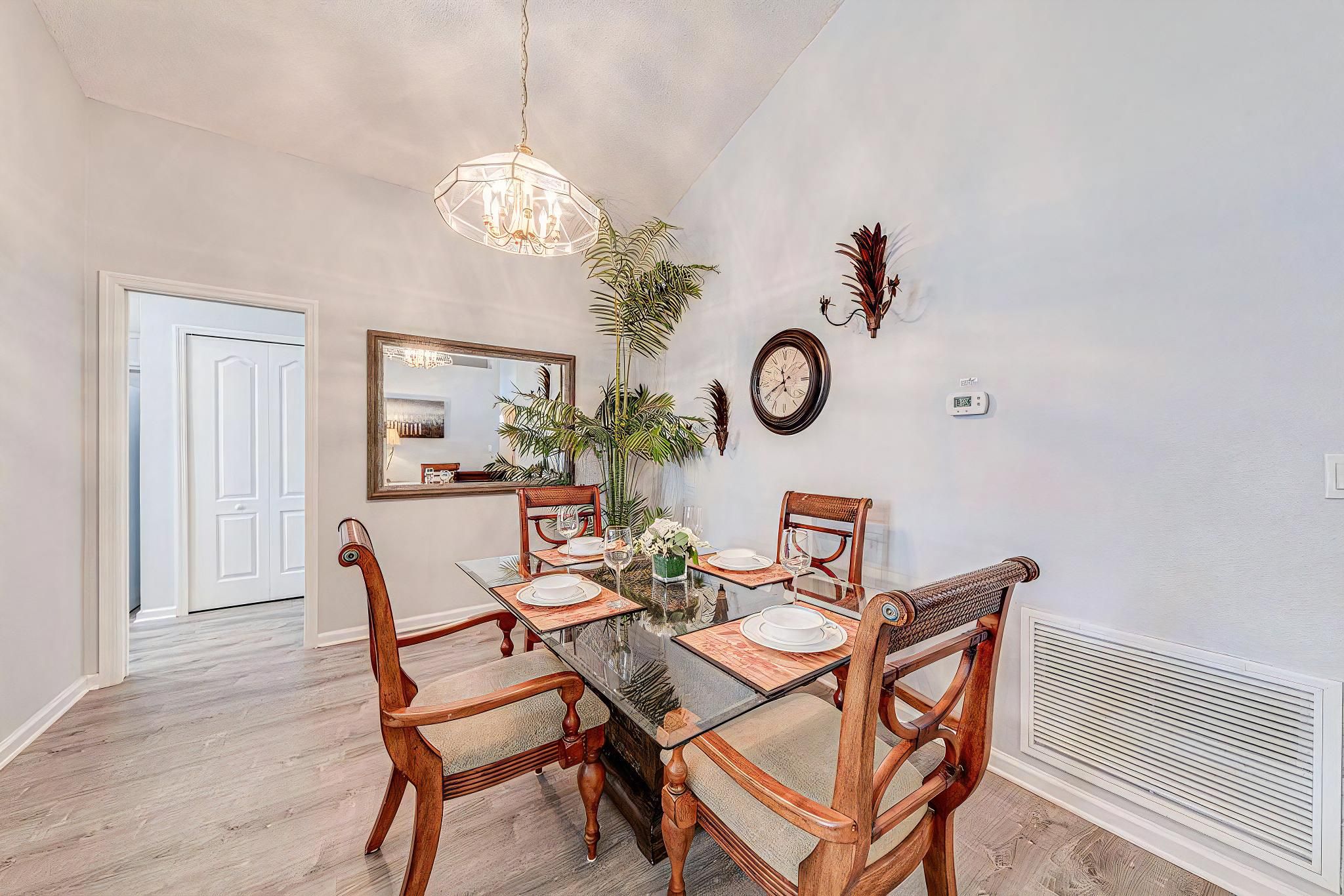 A dining area with a glass table, wooden chairs, a chandelier, a mirror, and wall-mounted decor.